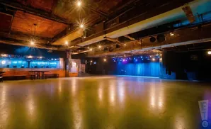 An empty, industrial concert venue with a stage, blue curtains, and a polished concrete floor reflecting stage lights.
