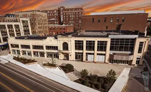 Aerial view of a light-colored building with large windows, flanked by older brick buildings under a dramatic sunset sky.