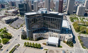 Aerial view of a large, glass-fronted building with a helipad on top, surrounded by city streets and green spaces.