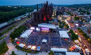 Aerial view of an outdoor concert and festival at dusk, with a stage, seating, tents, and food vendors near an industrial steel plant.