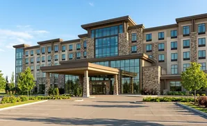 Exterior view of a modern, multi-story hotel with a large glass entrance and stone accents, surrounded by green trees and a parking lot.