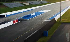 A drag strip with blue and white lane markers and grandstands in the distance.