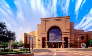 Exterior of a tan-colored building with a large arched glass entrance, under a bright blue sky with wispy clouds.