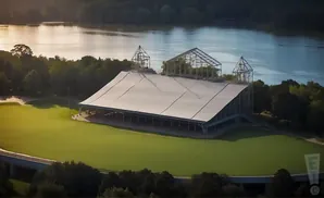 Aerial view of a large white tent structure with open sides on a green grassy field next to a body of water.