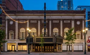 An old theater facade with a marquee, lit by strings of lights and street lamps, at dusk.