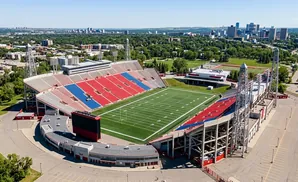 Aerial view of a stadium with red and blue seats, a green football field, and a city skyline in the distance.