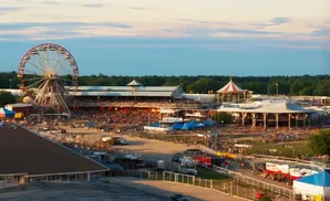 A Ferris wheel and grandstand are illuminated at a crowded fairgrounds during sunset.