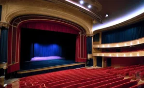 A grand theater with red seats, a stage with blue curtains, and ornate golden detailing.