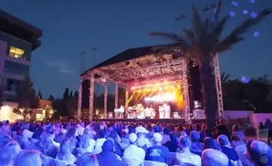 An outdoor concert at dusk with a band performing on a lit stage to an audience seated in chairs.