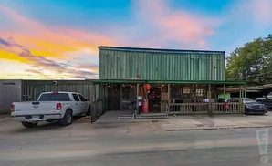 Exterior of a rustic, green wooden building with a covered porch, a white pickup truck parked to the left, and a dark car to the right, under a vibrant …