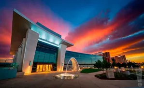 Modern building with glass facade and arched fountain at sunset with vibrant orange and blue sky.