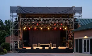 Outdoor stage with lights, speakers, and a dark curtain backdrop, with a brick building to the right.