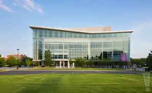 Modern glass building with a curved roof, surrounded by green grass and trees, under a blue sky.