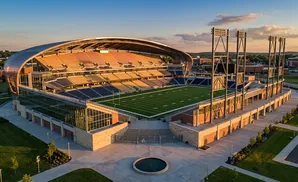 Aerial view of a modern football stadium with a large curved roof and colorful seating during a sunset.