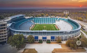 Aerial view of an empty football stadium at sunset, with a green field and teal seats.