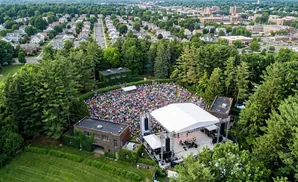 An aerial view of a large outdoor concert with a crowd gathered on grass and a band performing on stage.