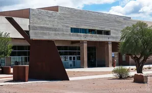 Modern building with concrete and rust-colored metal accents, featuring large windows and a tree.