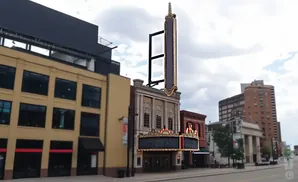 Marquee of an old theater with a tall vertical sign, next to a tan building and city skyline.