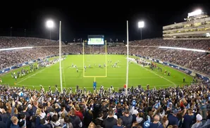 A panoramic view of a nighttime football game in a packed stadium, with bright lights illuminating the field and fans cheering.