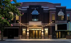 Exterior of a dark brick building with arched windows, string lights, and a marquee at dusk.