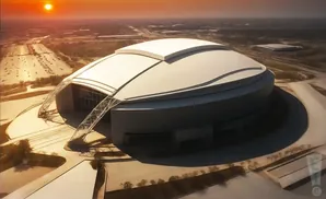 Aerial view of a modern domed stadium at sunset, with a sunlit parking lot and surrounding landscape.