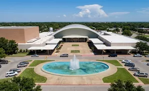 Aerial view of a large modern building with a domed roof, surrounded by trees, a fountain, and a parking lot with cars.