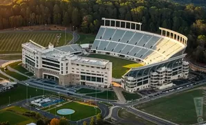 Aerial view of a modern stadium with lush green fields and trees, bathed in golden sunlight.