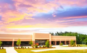 A modern building with large windows and a beige facade stands against a vibrant sunset sky with pink, orange, and purple clouds.