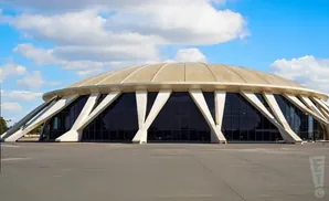 A domed building with triangular concrete supports and dark windows under a blue sky with clouds.