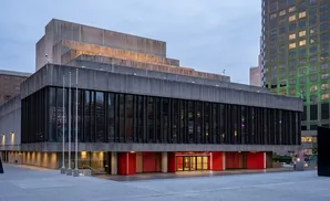 A modern concrete building with glass windows and red accents, set against a cloudy sky.