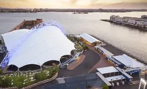 Aerial view of a waterfront venue with a large white tent structure and nearby buildings.