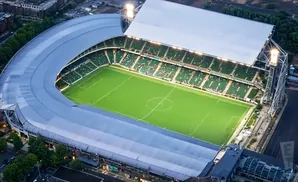 Aerial view of a green soccer stadium with a curved, silver roof and bright lights.