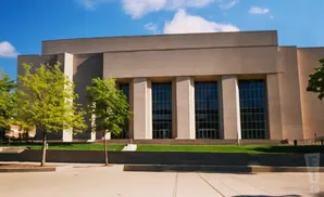 A large, tan building with tall windows and trees in front on a sunny day.