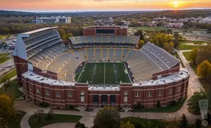 Aerial view of a brick stadium with a football field and yellow-seated stands at sunset.
