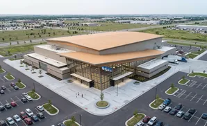 Aerial view of a modern building with large glass windows and a wooden roof, surrounded by parking lots and green spaces.