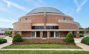 A circular brick building with a domed roof is surrounded by greenery and a blue sky.