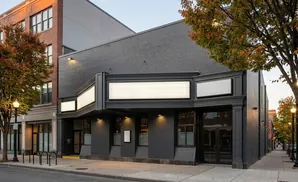A dark grey building with marquee signs and illuminated windows stands on a street with trees.