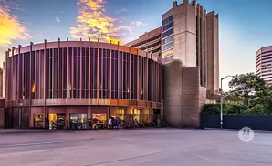 A circular building with vertical windows and a tall concrete building behind it at sunset.