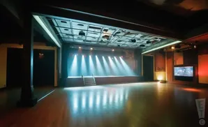 An empty, dimly lit theater stage with bright spotlights and a wide wooden floor.