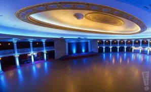 A grand ballroom with a stage, columns, and ornate ceiling, illuminated by blue lights reflecting off the floor.
