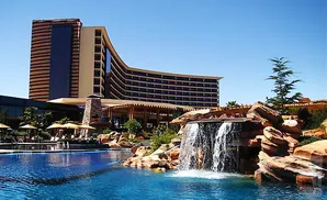 A modern hotel with a curved facade overlooks a swimming pool with a waterfall and rocky landscaping.