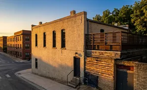 A historic brick building with windows, a fenced patio, and an alleyway under a clear sky at sunset.