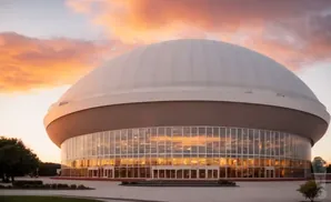 A domed stadium with a glass facade reflects an orange sunset.