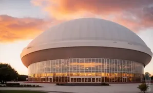 A domed stadium with glass walls at sunset, reflecting the warm sky.