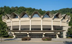 Brutalist concrete building with a jagged roofline and large glass windows against a backdrop of lush green hills.