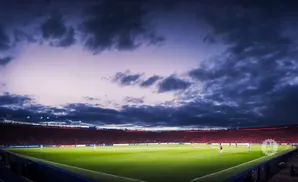Soccer stadium at dusk with a vibrant green field, a crowd in red, and dramatic clouds in the sky.