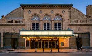 Facade of a vintage theater at dusk, with a lit marquee over the entrance and arched windows above.