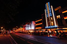 Neon blue Breakwater Hotel sign glowing at night in Miami South Beach above a lively, illuminated street and dining patio.