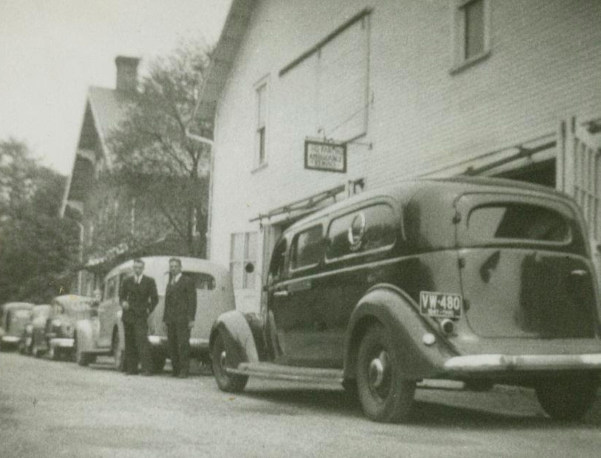 funeral directors Dick Snyder and Henry Hosler stand between cars 1937 Ford, 1940 LaSalle, 1946 Chrysler, and 1948 Buick outside of the Bellville Snyder Funeral Home