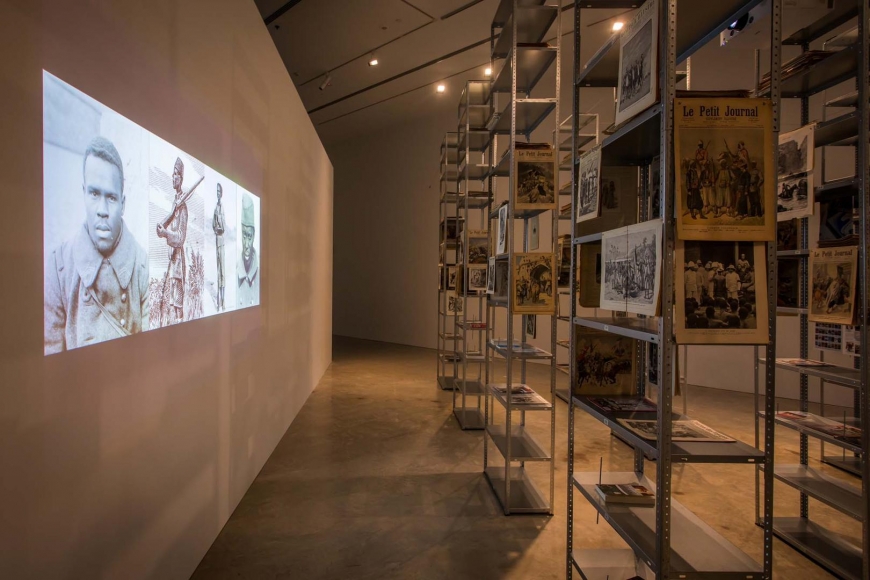 Kader Attia, installation view (left to right): The Debt, 2013, 2-channel digital video, projection, colour, based on the original 2-channel slide presentation; The Culture of Fear: An Invention of Evil, 2013, metal shelves, antique magazines, books, metal screws; Kader Attia, Museum of Contemporary Art Australia, Sydney, 2017, courtesy the artist, Galerie Nagel Draxler, Berlin/Cologne, Lehmann Maupin, New York and Hong Kong and Galleria Continua, San Gimignano, Les Moulins, Beijing, Habana. Image courtesy the artist and Museum of Contemporary Art Australia © the artist, photograph: Anna Kučera